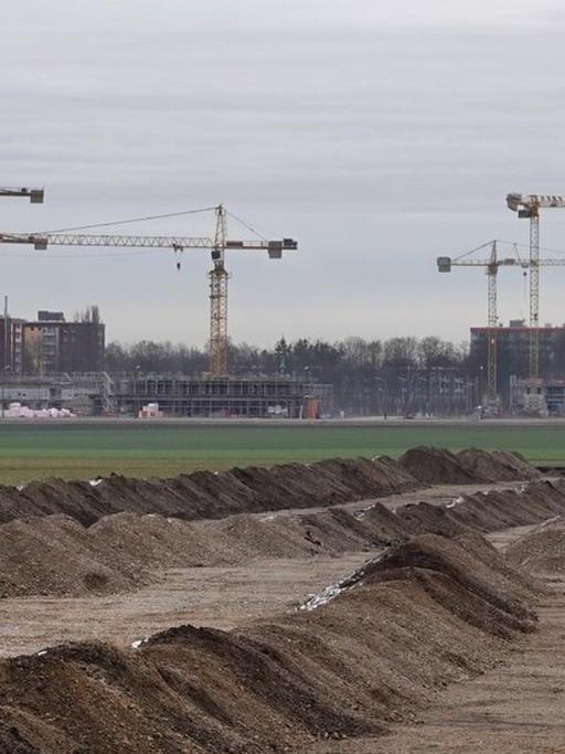 Hinter einer grünen Wiese an einer Großbaustelle im Münchner Stadtteil Freiham stehen Kräne. Auf der neuen Bebauungsfläche ist der Bodenaushub in Reihen angeordnet.