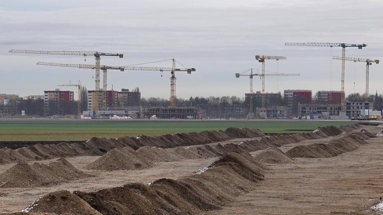 Hinter einer grünen Wiese an einer Großbaustelle im Münchner Stadtteil Freiham stehen Kräne. Auf der neuen Bebauungsfläche ist der Bodenaushub in Reihen angeordnet. Hinter einer grünen Wiese an einer Großbaustelle im Münchner Stadtteil Freiham stehen Kräne. Auf der neuen Bebauungsfläche ist der Bodenaushub in Reihen angeordnet.
