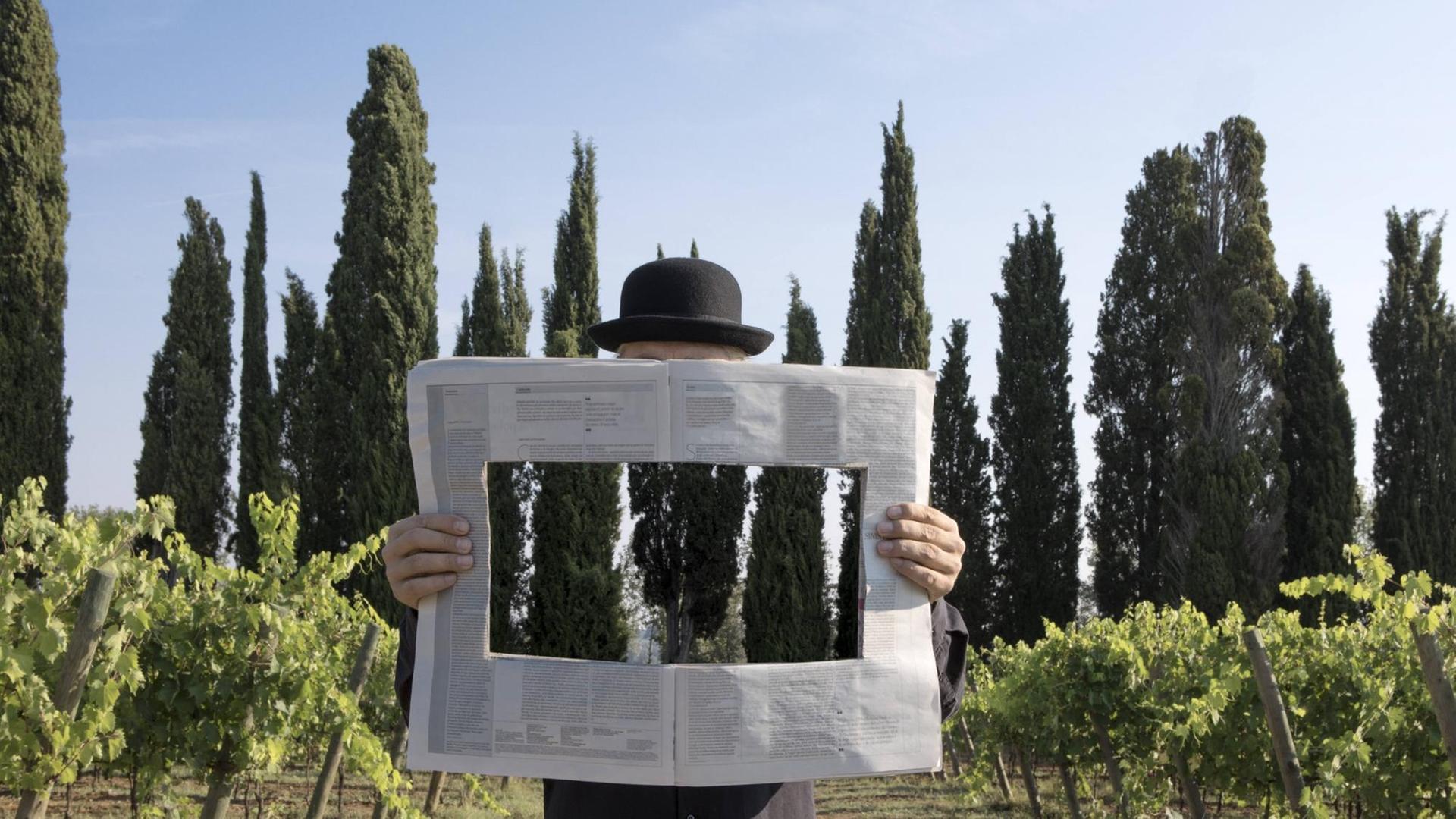 Ein Mann hält eine Zeitung in der Hand, die in der Mitte ein Loch hat, durch das man die toskanische Landschaft im Hintergrund sieht Ein Mann hält eine Zeitung in der Hand, die in der Mitte ein Loch hat, durch das man die toskanische Landschaft im Hintergrund sieht