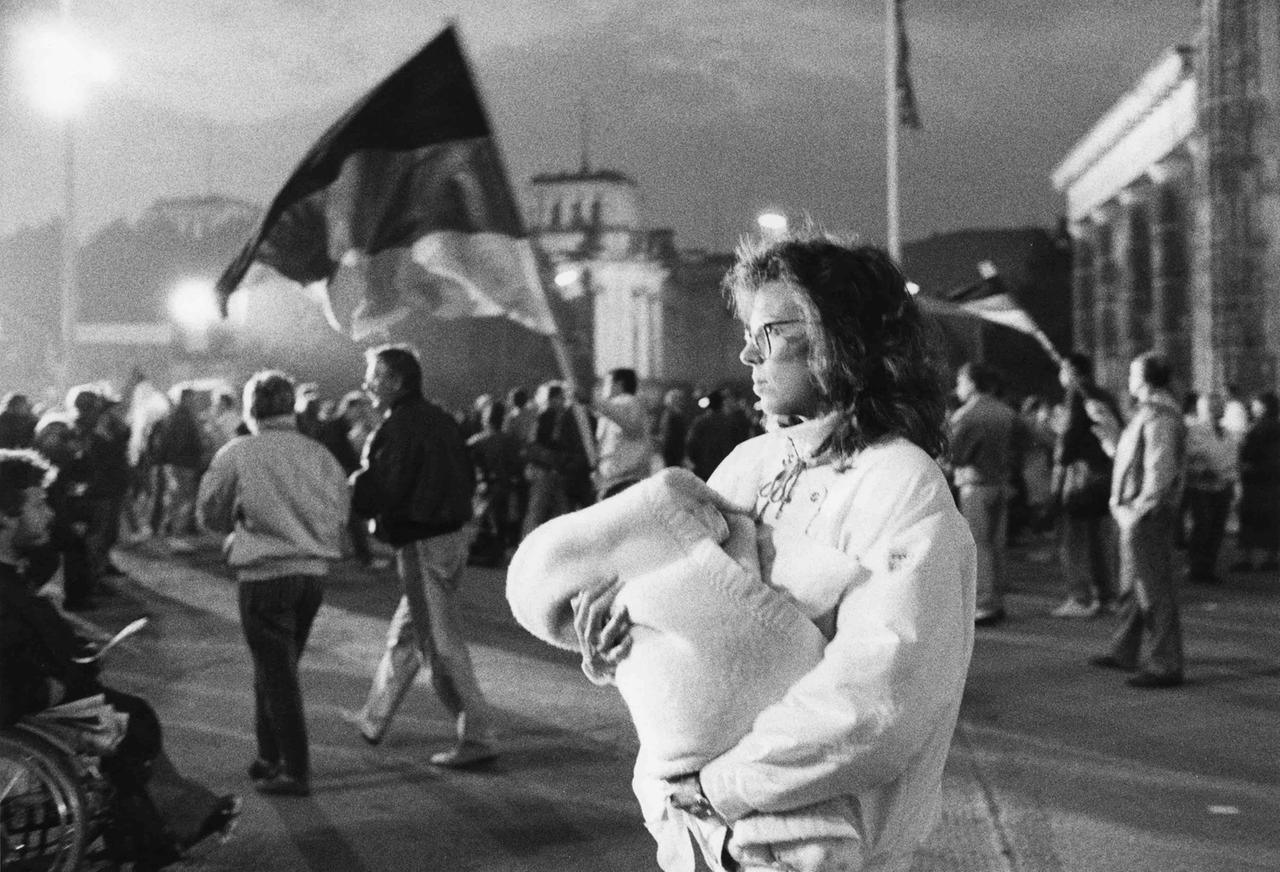 Das Schwarz-Weiß-Foto zeigt Menschen vor dem Brandenburger Tor in Berlin, im Vordergrund eine junge Frau, die einen Säugling im Arm hält.