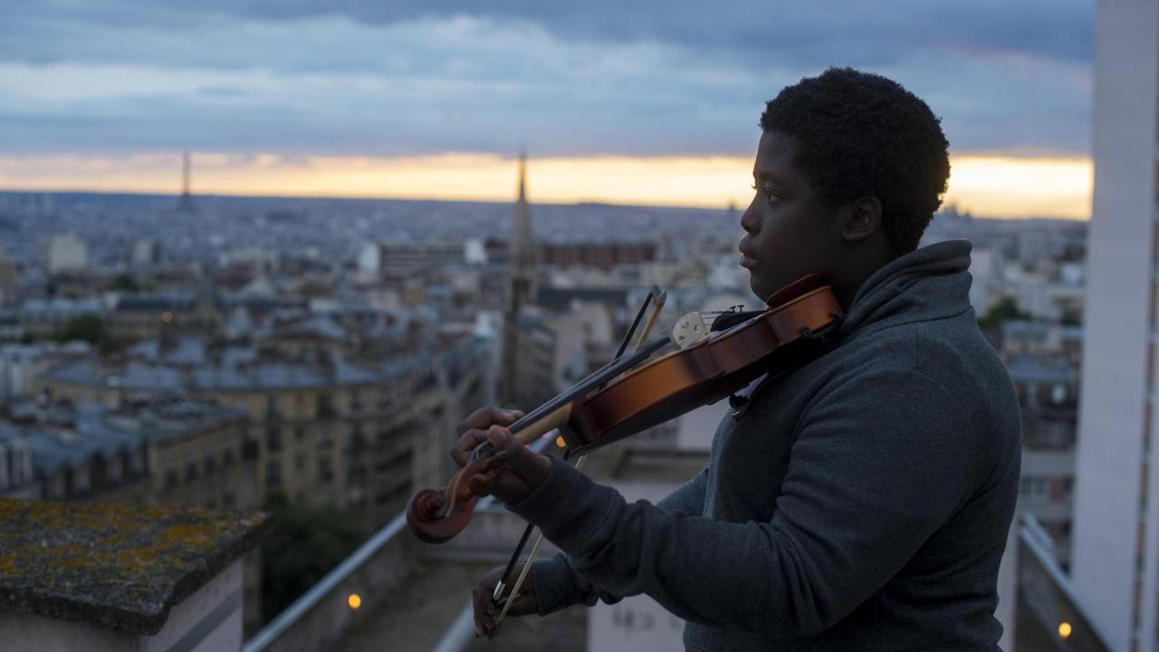 Szenenbild aus "La Mélodie - Der Klang von Paris". Ein Junge steht mit seiner Geige auf einem Hausdach, im Hintergrund die Szenerie von Paris