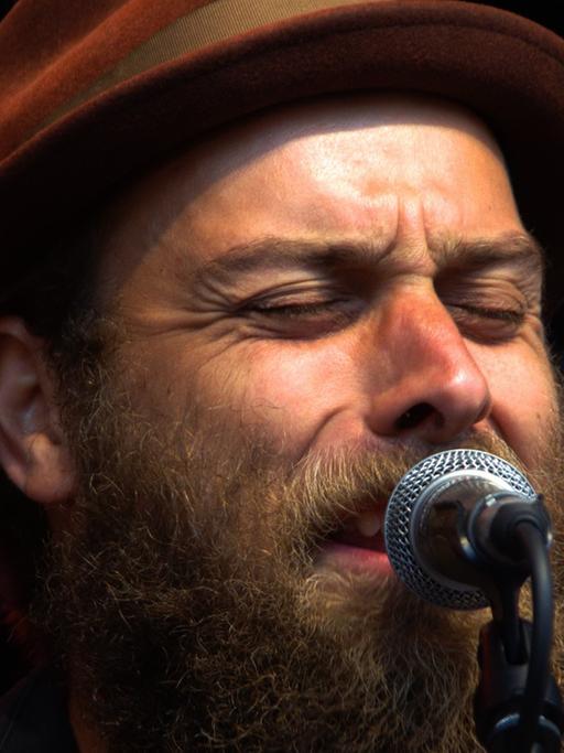 Ein Mann mit Hut und Vollbart steht mit geschlossenen Augen am Mikrofon und singt.
