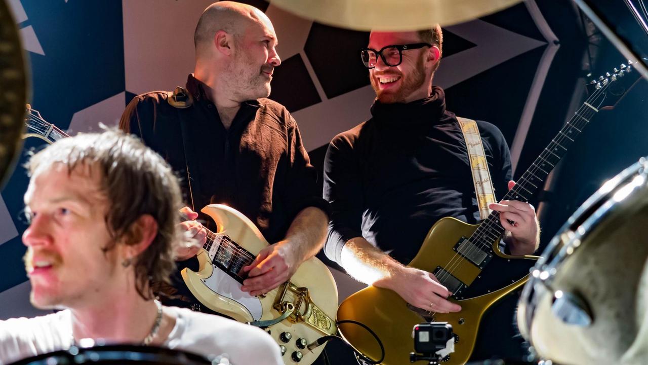 Ein Mann sitzt am Schlagzeug. Zwei Männer stehen mit Gitarren im Hintergrund. Ein Mann sitzt am Schlagzeug. Zwei Männer stehen mit Gitarren im Hintergrund.