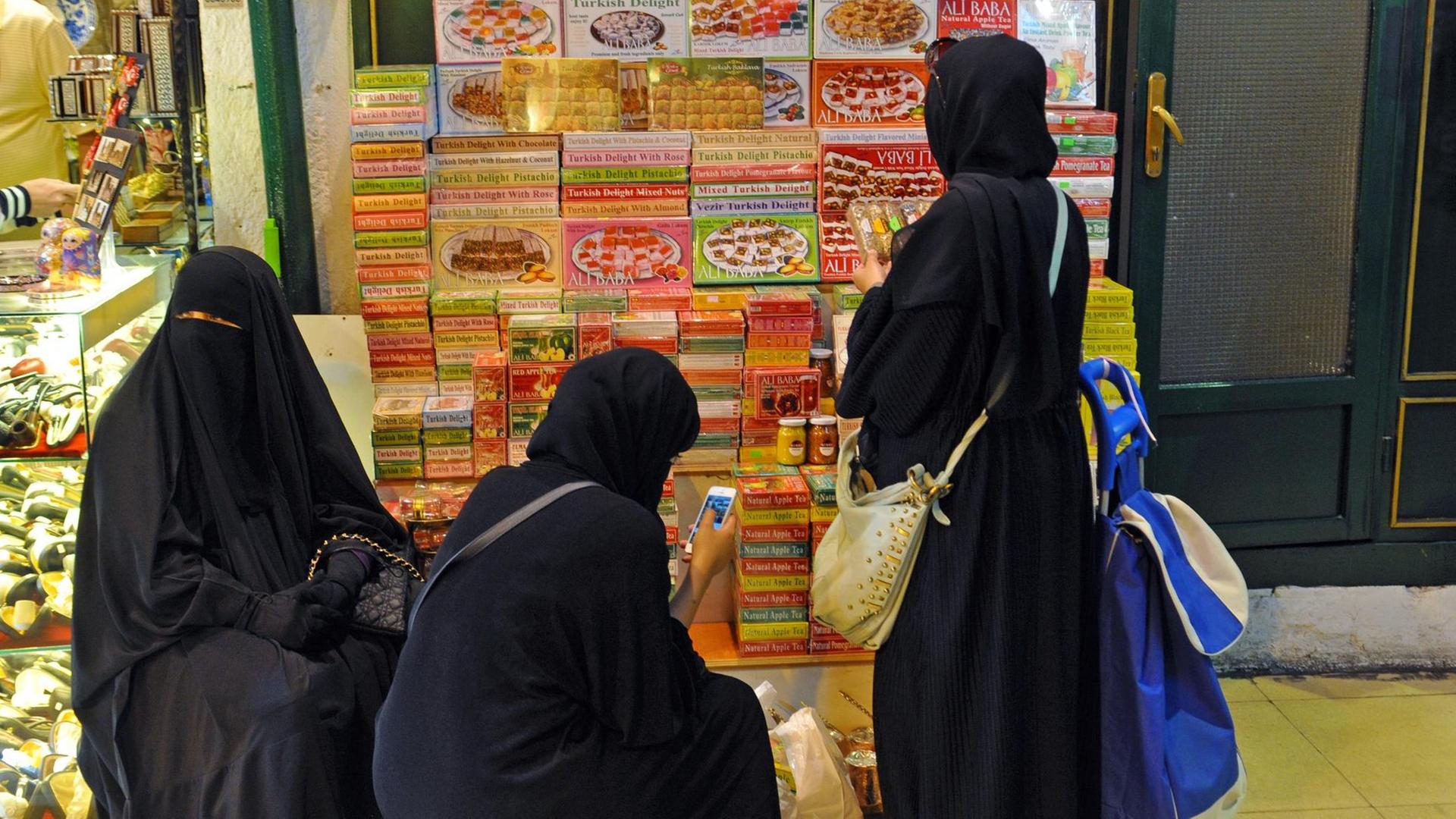Drei arabische Frauen aus den Golfstaaten in Vollverschleierung und Niqaab machen eine kleine Pause vor einem Stapel türkischer Süssigkeiten, aufgenommen im Großen Basar von Istanbul. Drei arabische Frauen aus den Golfstaaten in Vollverschleierung und Niqaab machen eine kleine Pause vor einem Stapel türkischer Süssigkeiten, aufgenommen im Großen Basar von Istanbul.