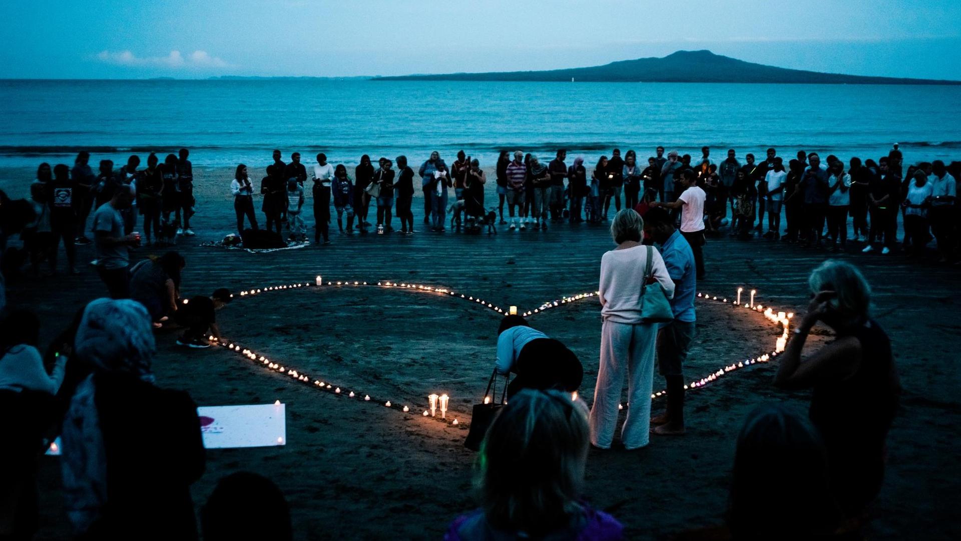 Eine Gruppe Menschen steht am Strand von Auckland in Neuseeland um ein Herz aus Kerzen. Eine Gruppe Menschen steht am Strand von Auckland in Neuseeland um ein Herz aus Kerzen.