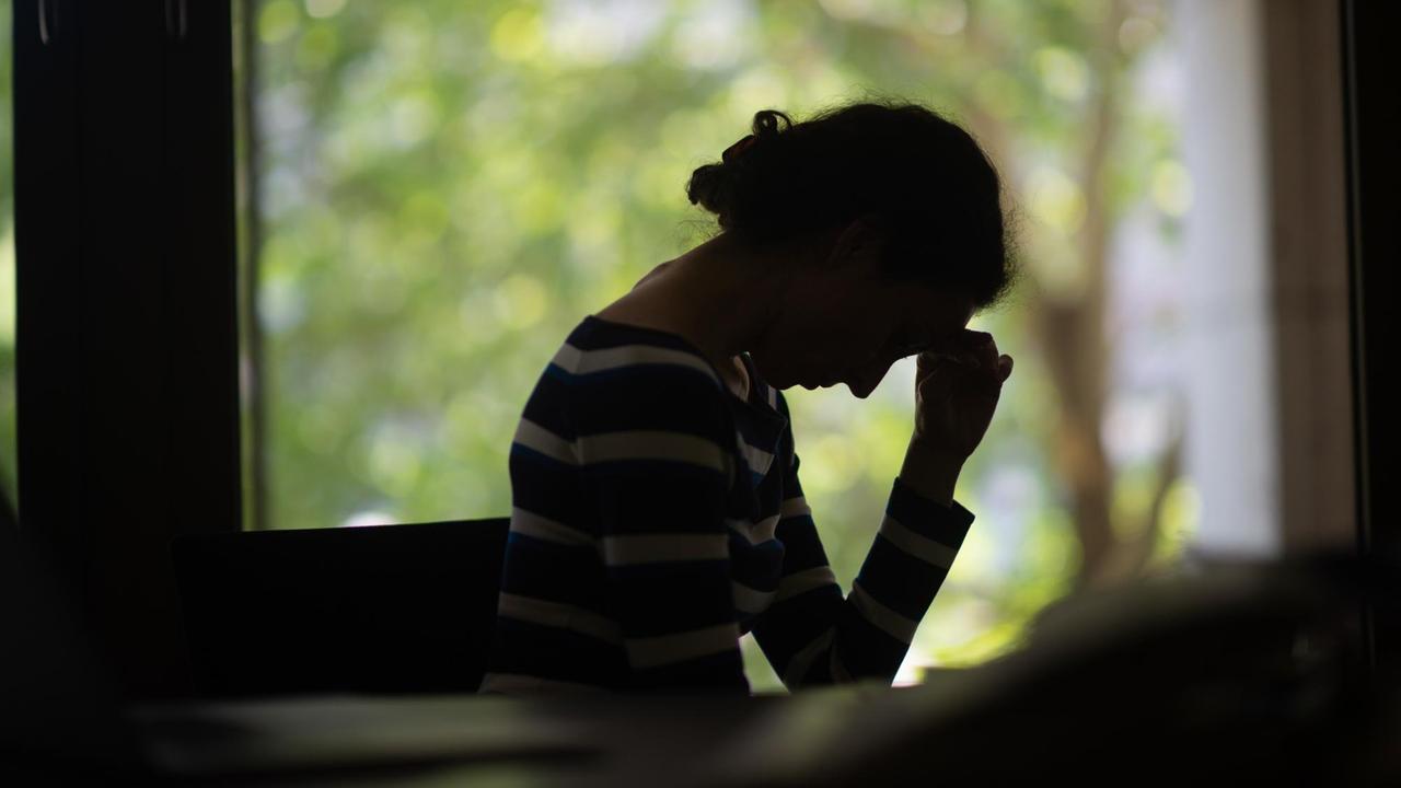 Die Silhouette einer Frau vor einem Fenster. Sie hat den Kopf in die Hand gestützt, ihre Haltung wirkt eingefallen. Die Silhouette einer Frau vor einem Fenster. Sie hat den Kopf in die Hand gestützt, ihre Haltung wirkt eingefallen.