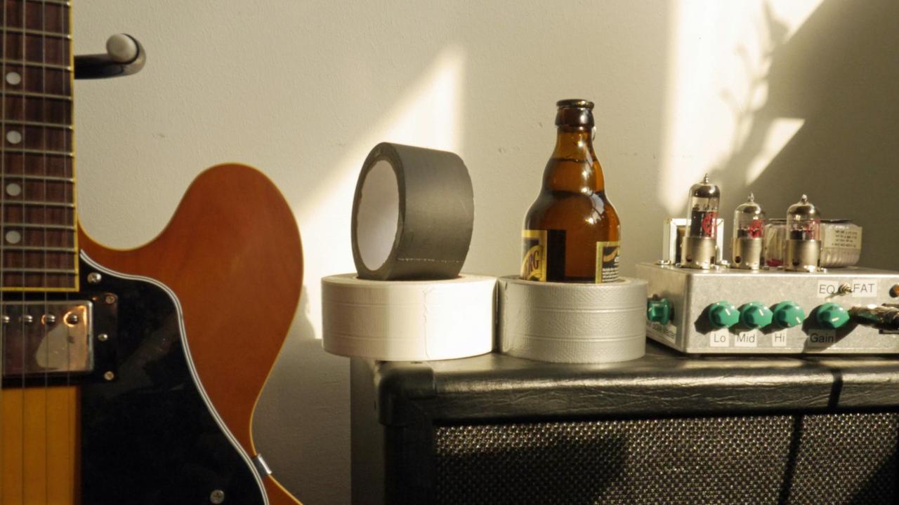 Drei Ollen Gewebeband stehen auf einem schwarzen Gitarrenverstärker, links eine Gitarre. Drei Ollen Gewebeband stehen auf einem schwarzen Gitarrenverstärker, links eine Gitarre.
