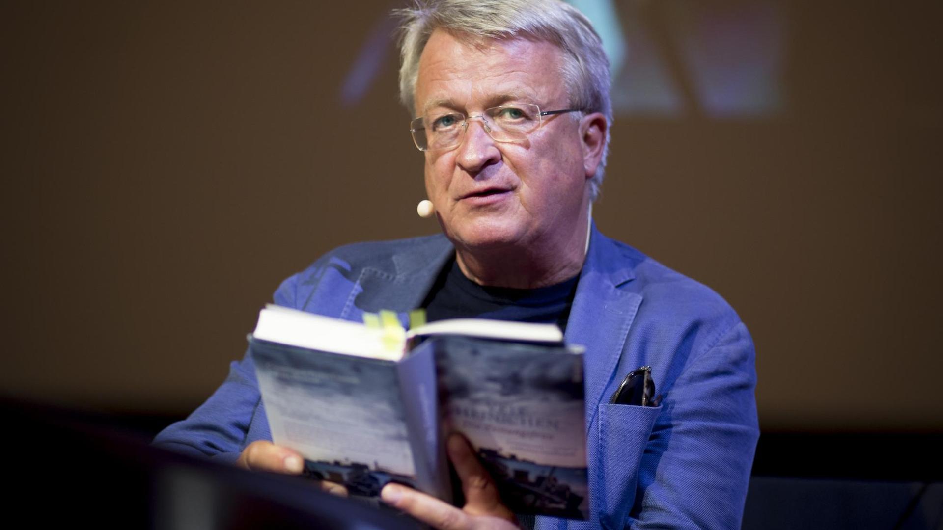 Der Autor Veit Heinichen sitzt auf einer Bühne und liest aus seinem Buch. Der Autor Veit Heinichen sitzt auf einer Bühne und liest aus seinem Buch.