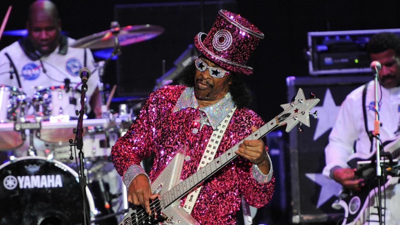 Bootsy Collins trägt auf der Bühne einen pink-roten Glimmeranzug und einen passenden Hut. Er hat eine Sonnenbrille mit Sternen drauf auf und spielt einen E-Bass, der Korpus ist sternförmig.