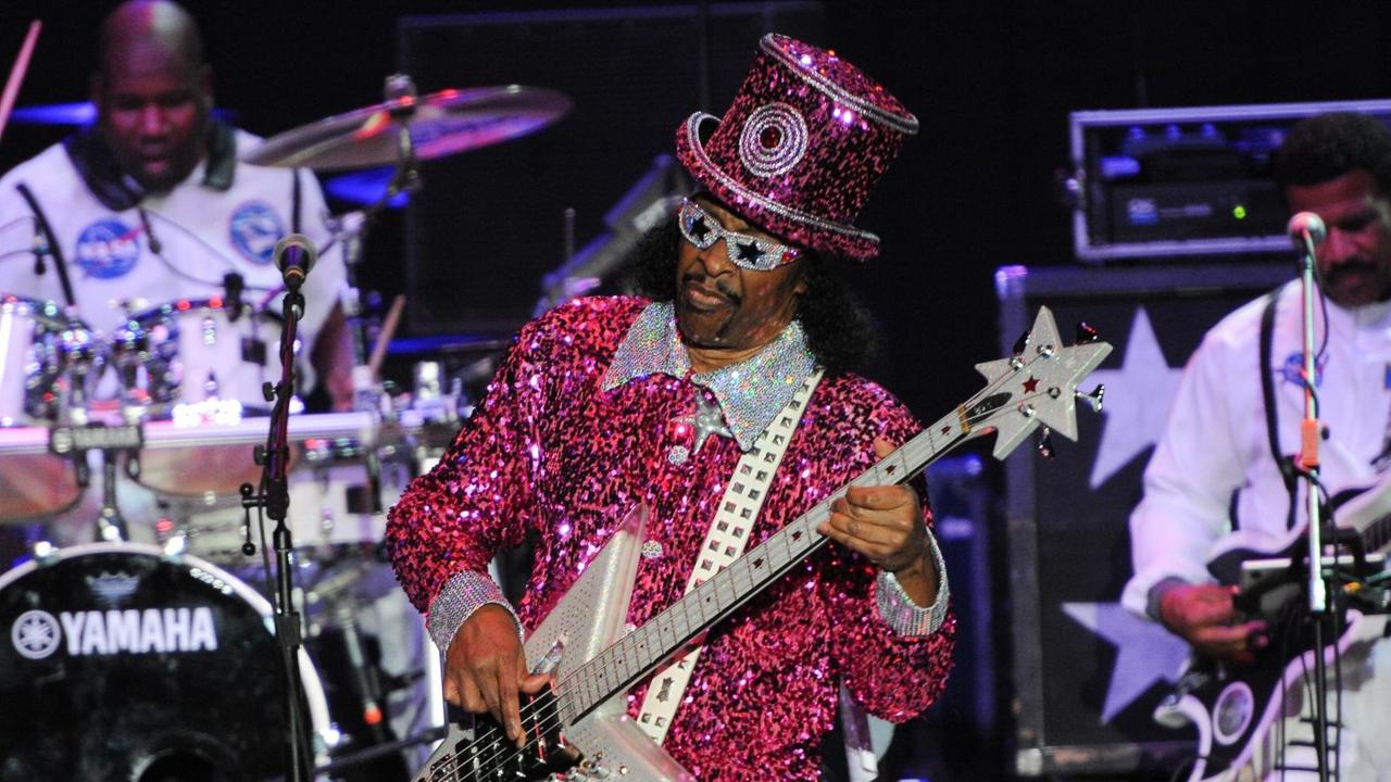 Bootsy Collins trägt auf der Bühne einen pink-roten Glimmeranzug und einen passenden Hut. Er hat eine Sonnenbrille mit Sternen drauf auf und spielt einen E-Bass, der Korpus ist sternförmig. Bootsy Collins trägt auf der Bühne einen pink-roten Glimmeranzug und einen passenden Hut. Er hat eine Sonnenbrille mit Sternen drauf auf und spielt einen E-Bass, der Korpus ist sternförmig.