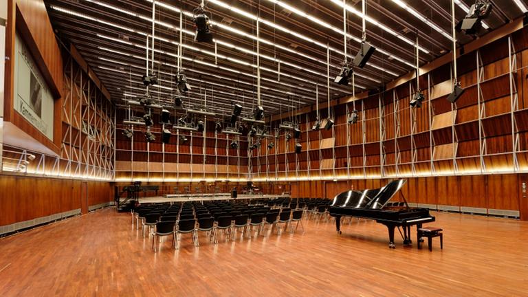 Zu sehen ist der menschenleere Kammermusiksaal im Deutschlandfunk-Gebäude. Ein großer Raum mit Parkett ausgelegt, darauf Stuhlreihen und davor ein Konzertpiano. Von der Decke hängen schwarze Scheinwerfer. Zu sehen ist der menschenleere Kammermusiksaal im Deutschlandfunk-Gebäude. Ein großer Raum mit Parkett ausgelegt, darauf Stuhlreihen und davor ein Konzertpiano. Von der Decke hängen schwarze Scheinwerfer.