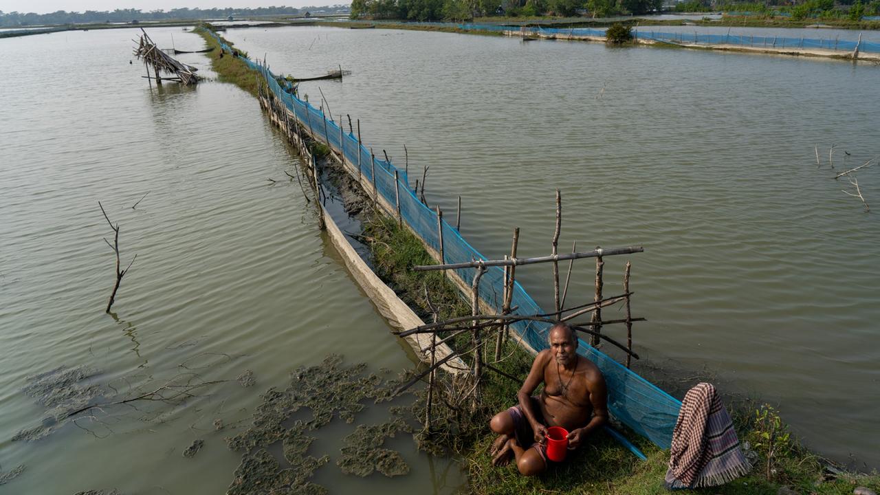 Shrimp-Farmer Ananda Biswas sitzt vor einem ins Wasser hineinragenden Steg mit Netzen an der Seite