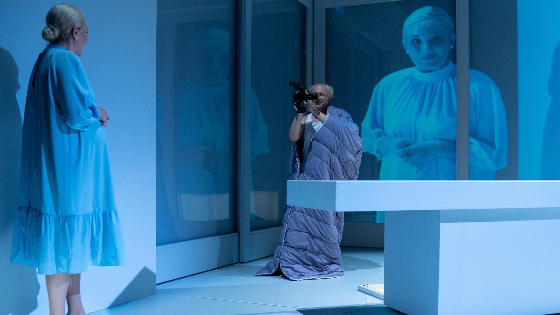 Auf einer hellblau ausgeleuchteten Theaterbühne filmt ein Mann eine Frau aus gewisser Entfernung. Ihr Bild wird im Hintergrund an die Wand projiziert.