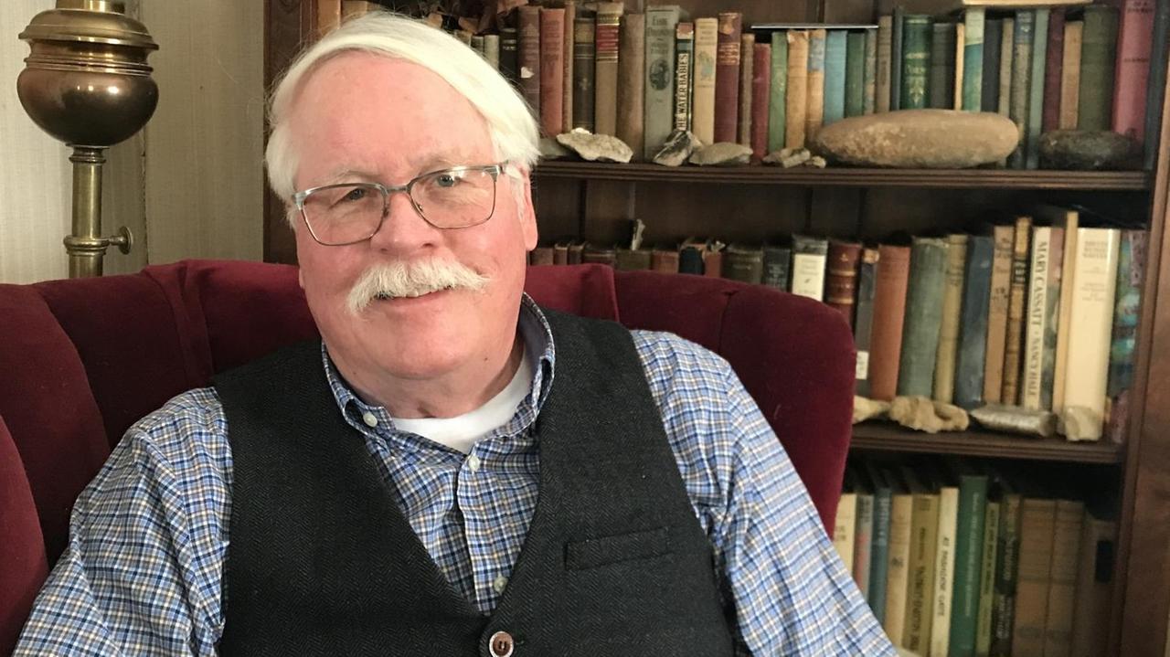 Grauhhariger Mann mit Bart in einem roten Sessel vor einem Bücherregal Grauhhariger Mann mit Bart in einem roten Sessel vor einem Bücherregal