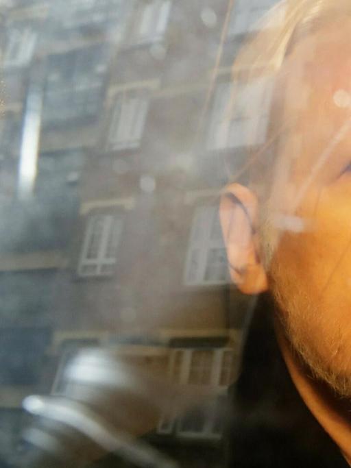 Julian Assange blickt mit gestutztem Bart und streng frisierten Haaren aus einem Busfenster, in dem Gebäude reflektieren.