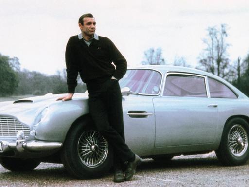 Sean Connery lässig an die Motorhaube eines Aston Martin DB5 gelehnt im Bond-Film Goldfinger aus dem Jahr 1964.