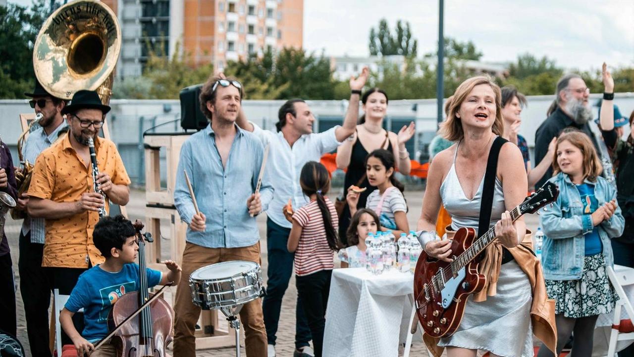 Eine Frau im hellen Sommerkleid steht in einem Kreis von Menschen, die Singen und verschiedene Instrumente spielen, während sie Gitarre spielt und laut singt. Eine Frau im hellen Sommerkleid steht in einem Kreis von Menschen, die Singen und verschiedene Instrumente spielen, während sie Gitarre spielt und laut singt.