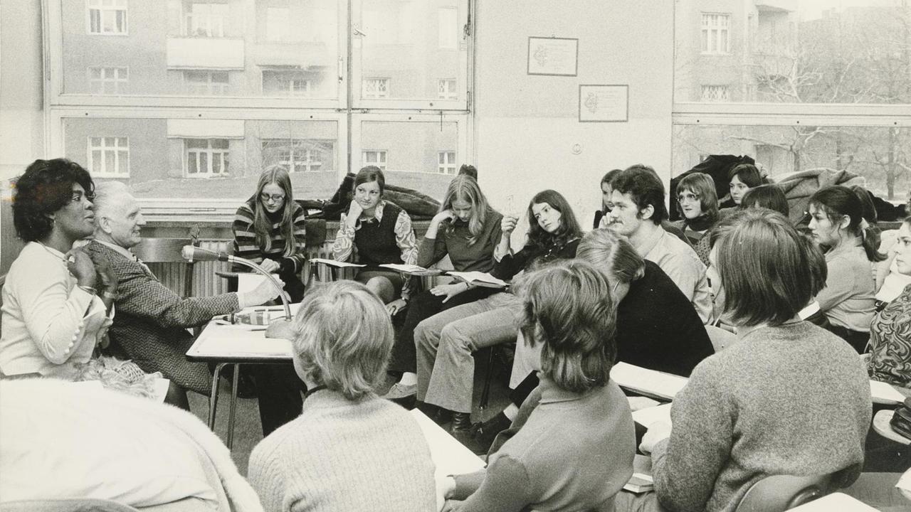Die Schwarz-weiß-Aufnahme zeigt Vera Little in einem Klassenzimmer. In einem Halbkreis sitzen die Schüler der Klasse vor ihr. Die Schwarz-weiß-Aufnahme zeigt Vera Little in einem Klassenzimmer. In einem Halbkreis sitzen die Schüler der Klasse vor ihr.