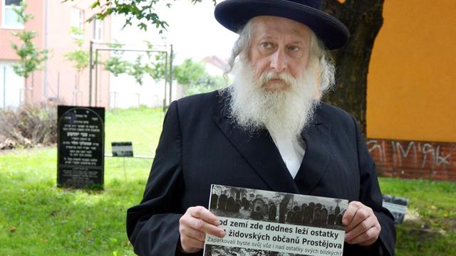 Ein Rabbiner auf dem Gelände des ehemaligen jüdischen Friedhofs in Prostějov hält eine alte Fotografie des Friedhofs mit zahlreichen Grabsteinen, die dort ursprünglich dicht nebeneinander standen.