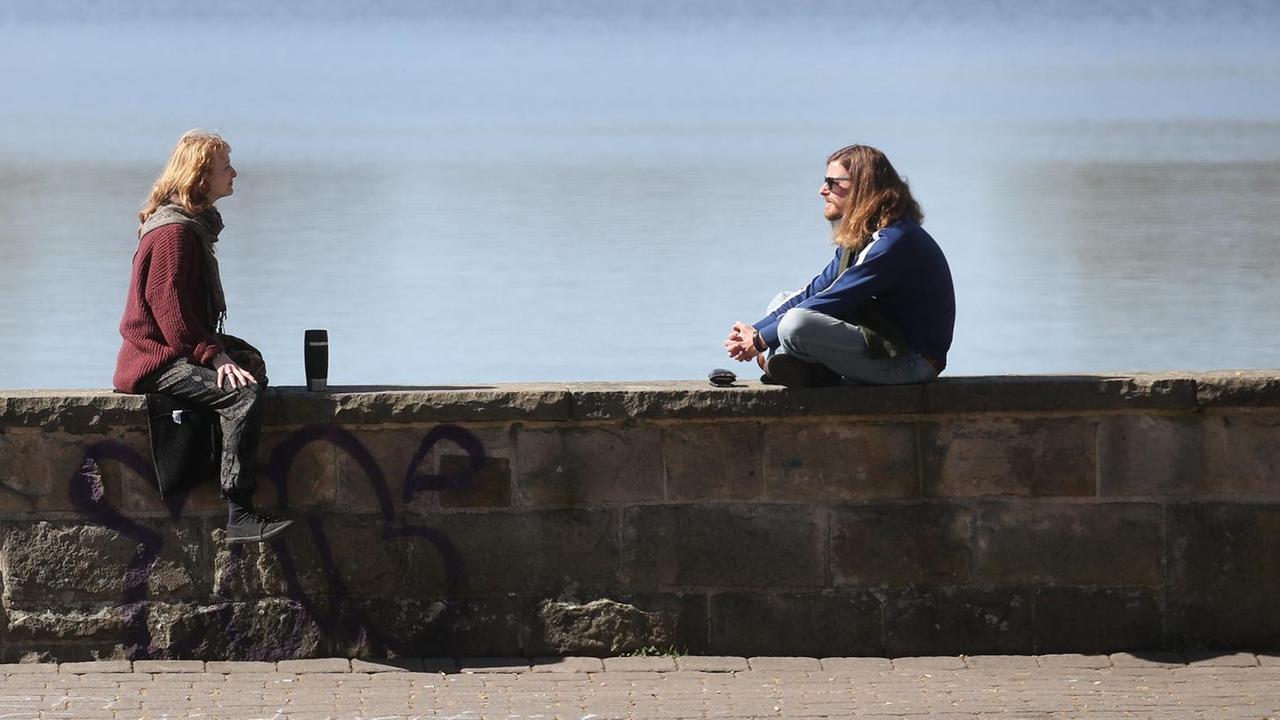 Zwei Menschen sitzen mit großem Abstand auf einer Mauer und unterhalten sich. Zwei Menschen sitzen mit großem Abstand auf einer Mauer und unterhalten sich.
