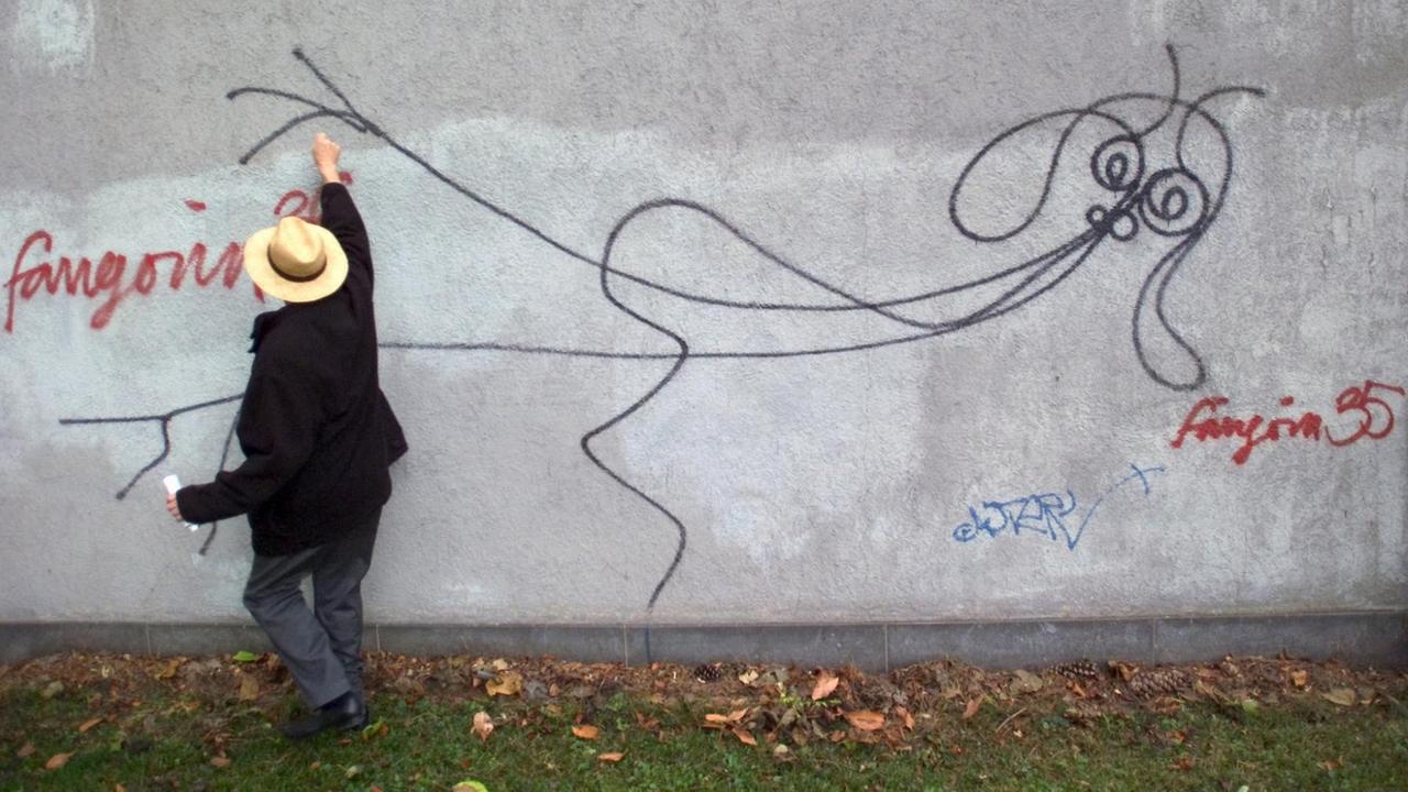 Ein Mann mit Hut steht vor einem Strichmännchchen an einer grauen Wand. Ein Mann mit Hut steht vor einem Strichmännchchen an einer grauen Wand.