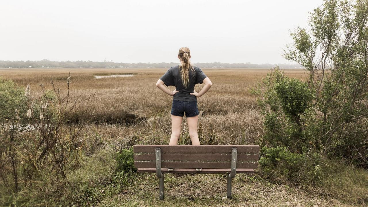 Die Rückansicht eines jungen Teenagermädchens, sie steht auf einer Bank und schaut über Felder in eine herbstliche Landschaft. (Symbolbild)