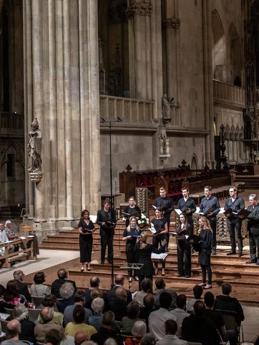 Die ORA Singers werden von Suzi Digby geleitet und gründeten sich 2016 und sind in London beheimatet. Hier sind sie im Halbrund aufgestellt im gotischen Regensburger Dom, die Sängerinnen und Sänger tragen schwarze Konzertkleidung, sie stehen vor dem eigentlichen Altarraum