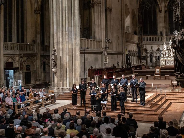Die ORA Singers werden von Suzi Digby geleitet und gründeten sich 2016 und sind in London beheimatet. Hier sind sie im Halbrund aufgestellt im gotischen Regensburger Dom, die Sängerinnen und Sänger tragen schwarze Konzertkleidung, sie stehen vor dem eigentlichen Altarraum