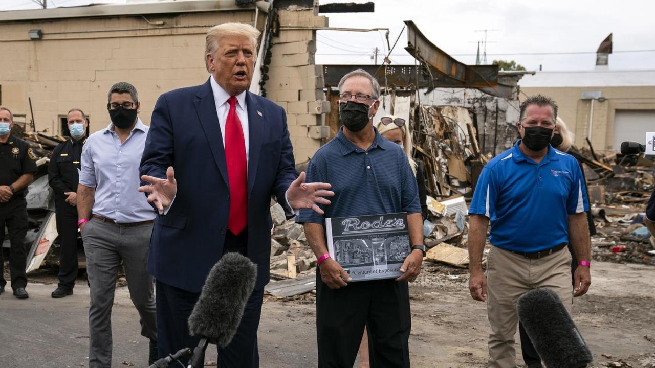 Donald Trump steht mit Anzug und roter Krawatte vor einem Mikrofon und hinter zerstörten Häusern. Donald Trump steht mit Anzug und roter Krawatte vor einem Mikrofon und hinter zerstörten Häusern.