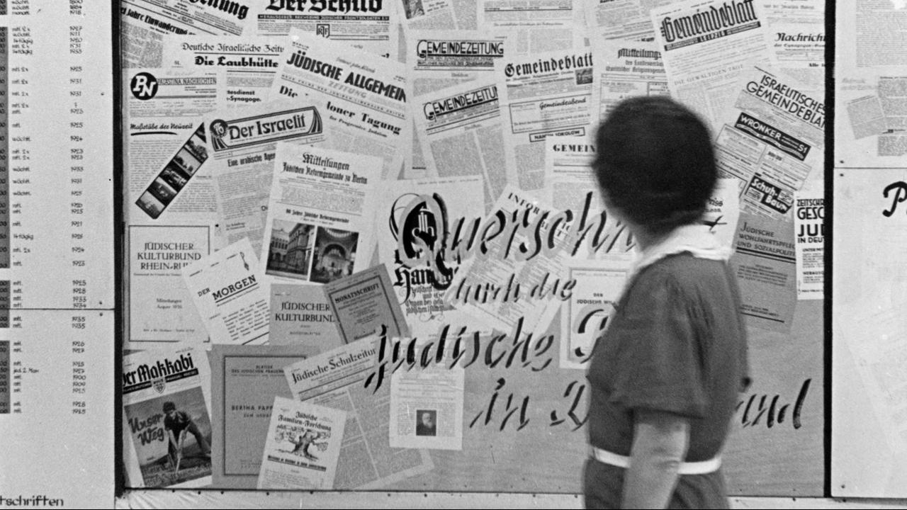 Eine Frau steht vor einer Wand mit verschiedenen Zeitungs-Titelseiten.