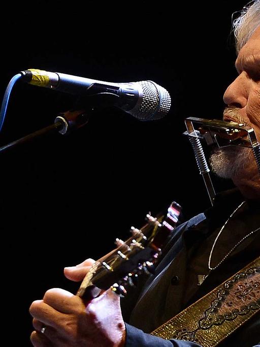 Der Musiker steht auf einer Bühne, wobei er vor einem Mikrophon steht, seine Gitarre hält und gerade seine Mundharmonika bläst, die in einer Metallklemme steckt - eine Halterung um den Hals, für das freihändige Spielen.
