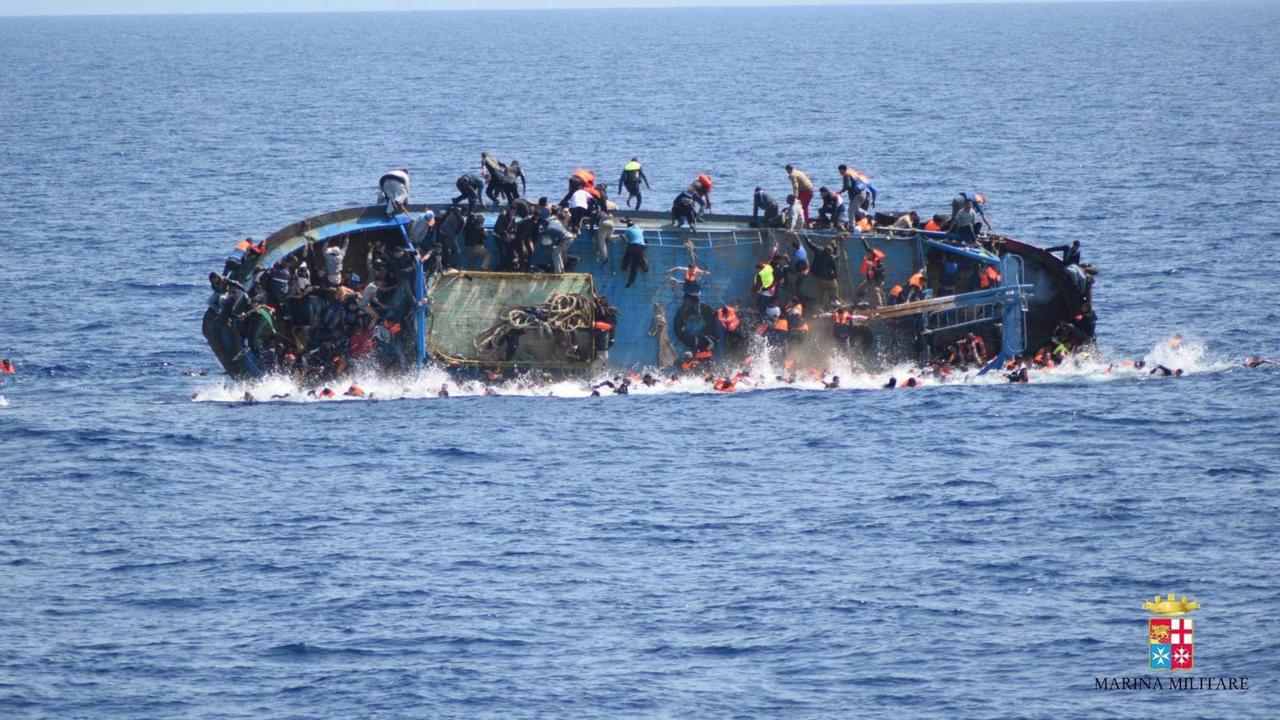 Ein Boot mit Flüchtlingen steht auf der Seite im Wasser, die Menschen fallen und springen zum Teil vom Boot ins Wasser. Ein Boot mit Flüchtlingen steht auf der Seite im Wasser, die Menschen fallen und springen zum Teil vom Boot ins Wasser.