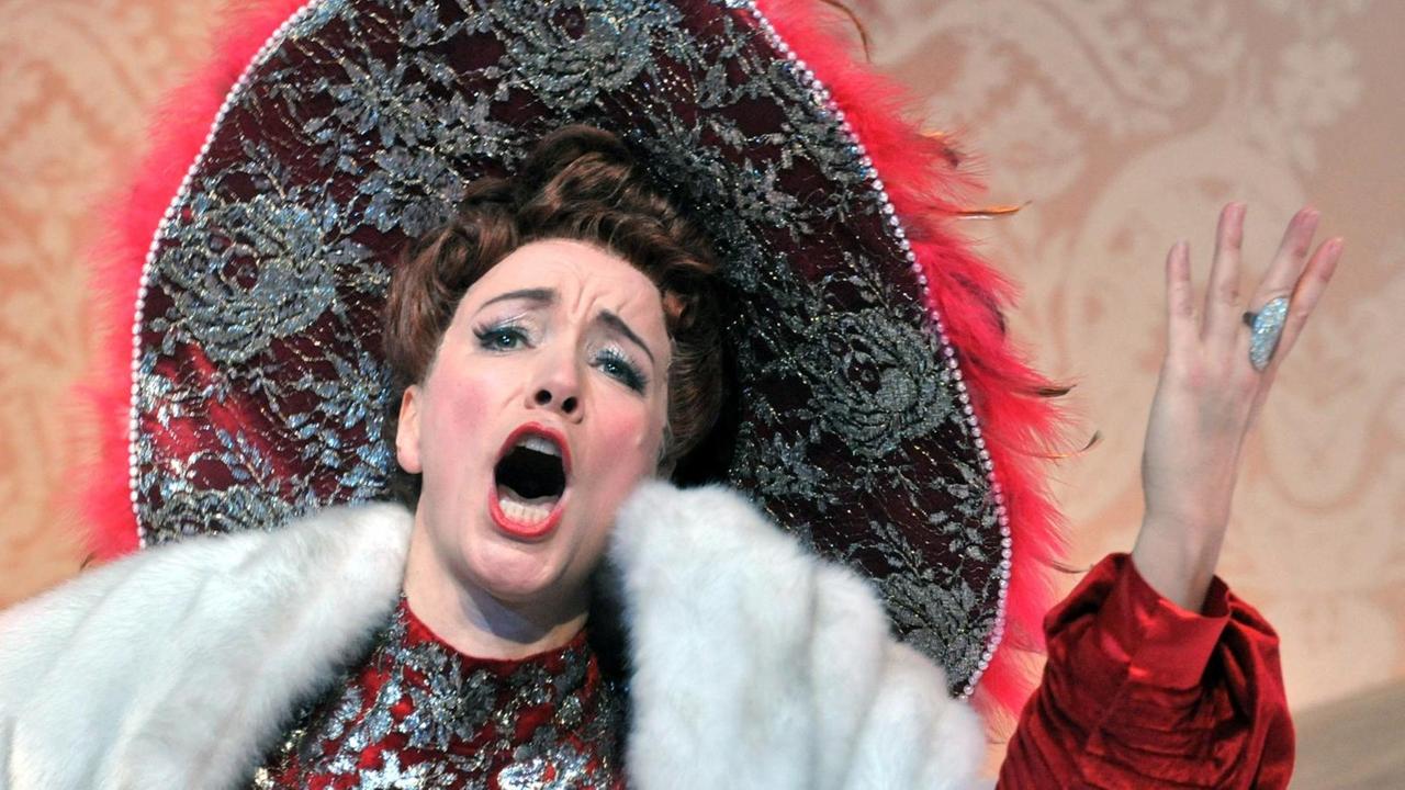 Eine Dame mit großem Hut, rotem Kleid und weißem Pelz steht singend auf einer Bühne. Eine Dame mit großem Hut, rotem Kleid und weißem Pelz steht singend auf einer Bühne.