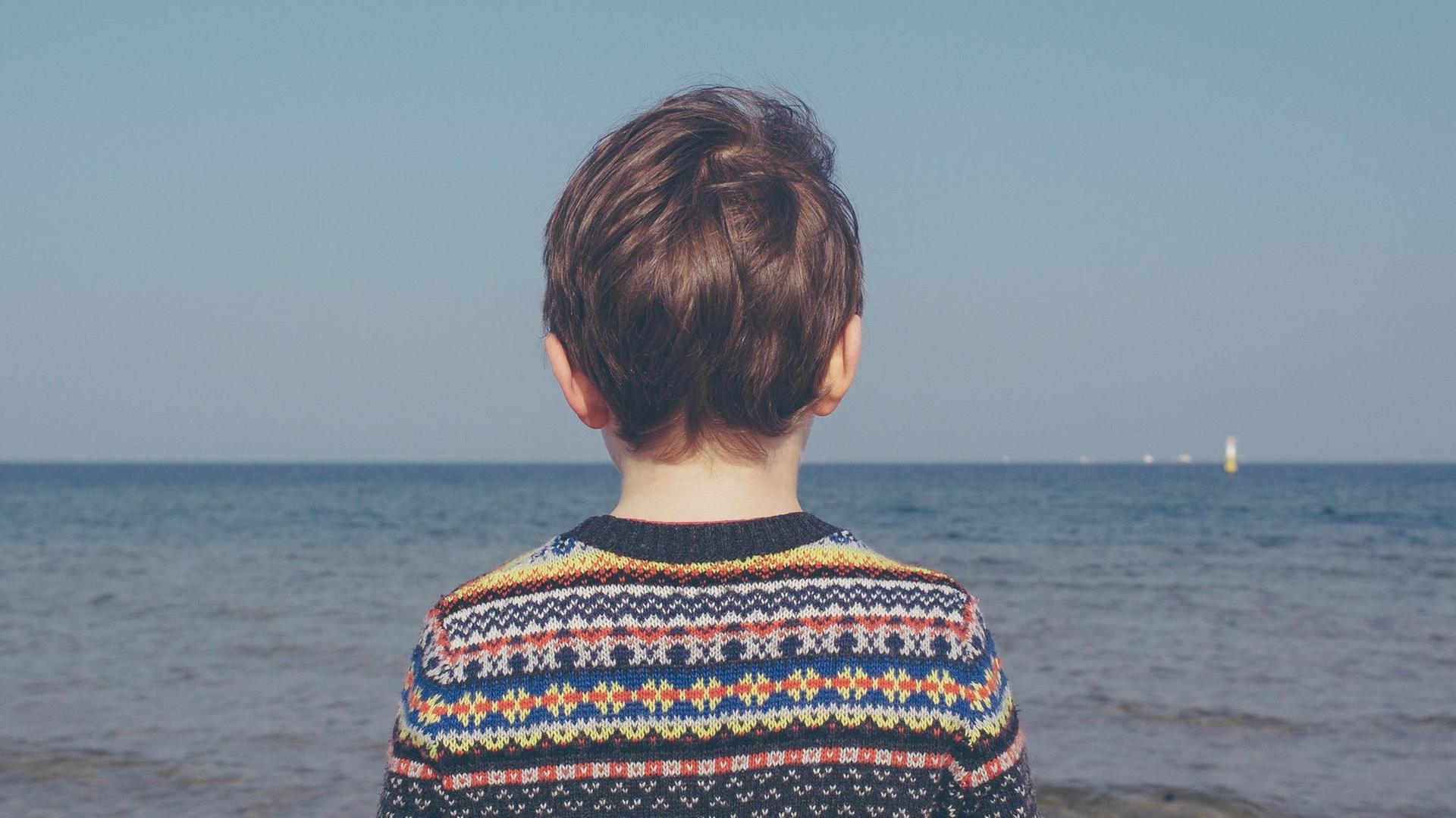 Ein Junge blickt alleine in Richtung Meer, bekleidet mit einem bunten Strickpulli. (Symbolbild)