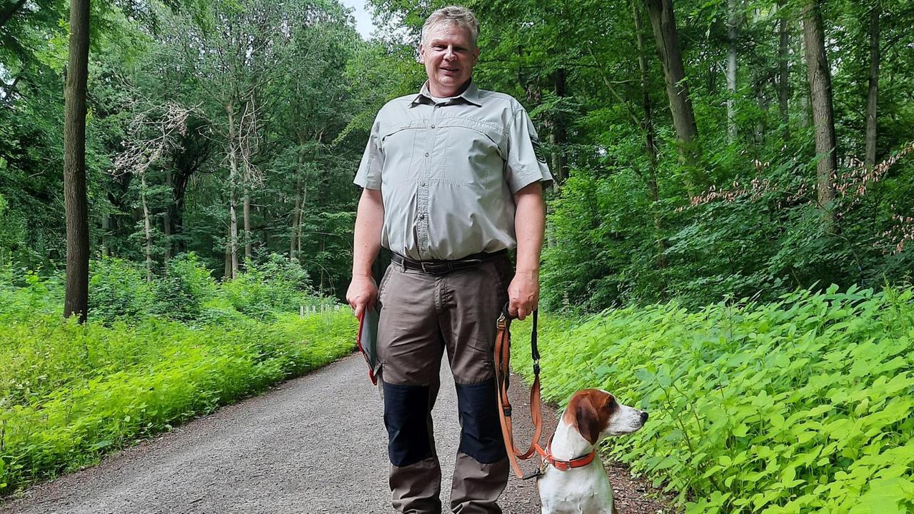 Ein älterer Mann steht auf einem Waldweg und hat einen Hund an der Leine.