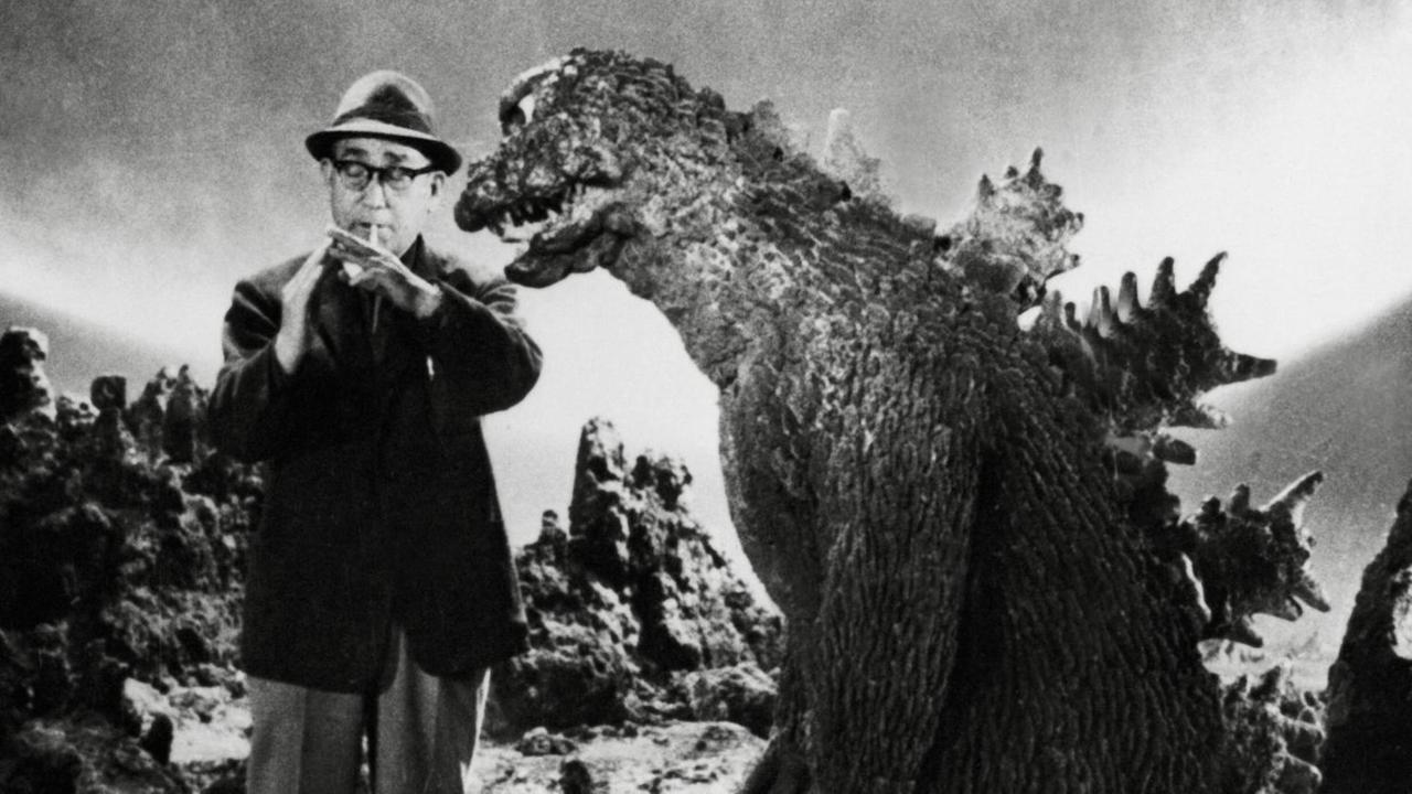 Schwarz-Weiß-Foto von Ishiro Honda, der sich eine Zigarette anzündet. Neben ihm steht der Godzilla-Darsteller in voller Montur, wodurch sich ein eigentümlicher Eindruck ergibt.