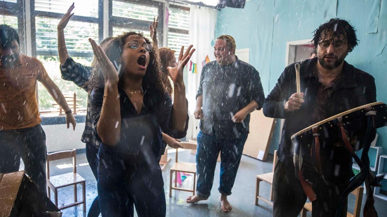 Auf dem Bild ist eine Szene aus dem Film "Andere Eltern" zu sehen. Menschen musizieren in einem Klassenzimmer. Es scheint von der Decke zu regnen.