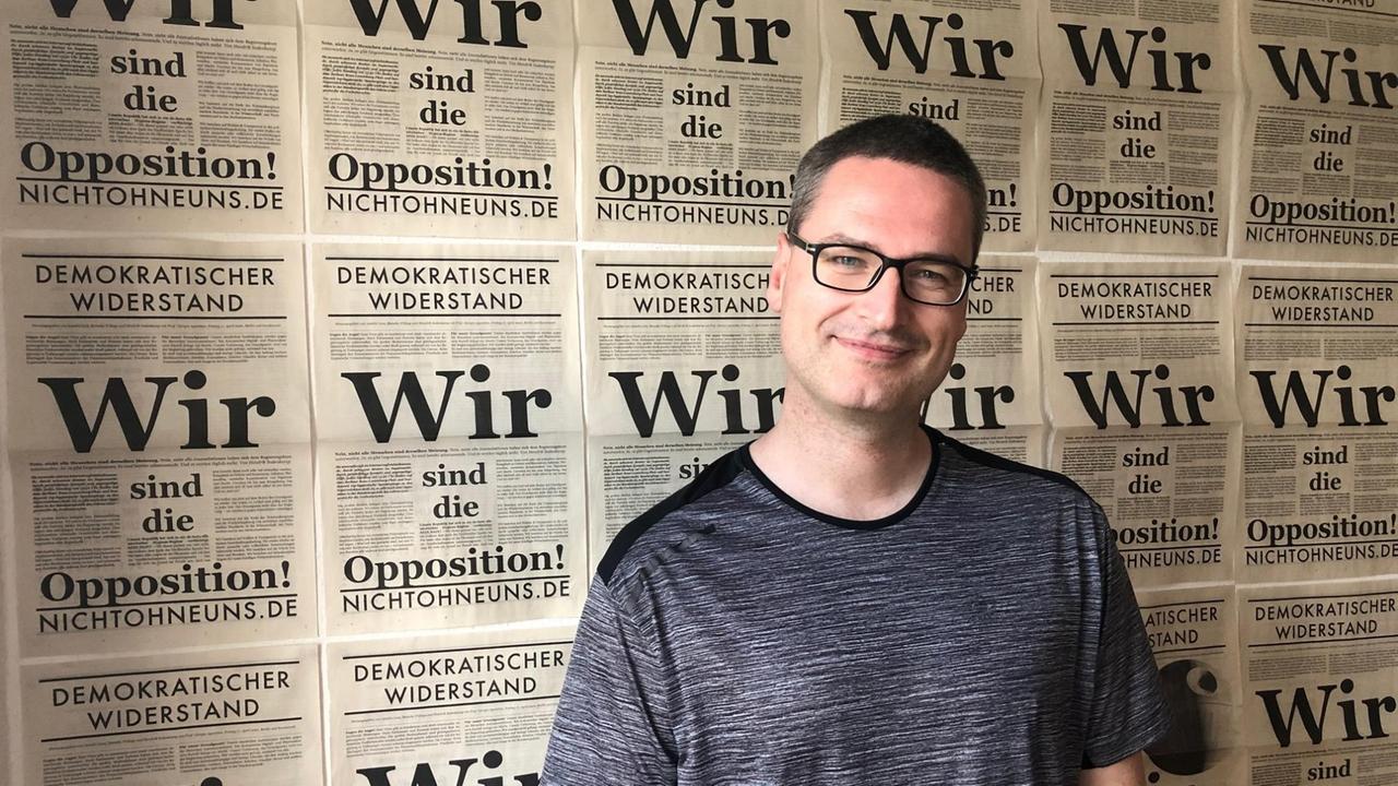 Ein Mann mit T-Shirt steh vor einer Wand mit den Titelseiten der Protestzeitung "Demokratischer Widerstand", auf denen zu lesen ist: "Wir sind die Opposition."