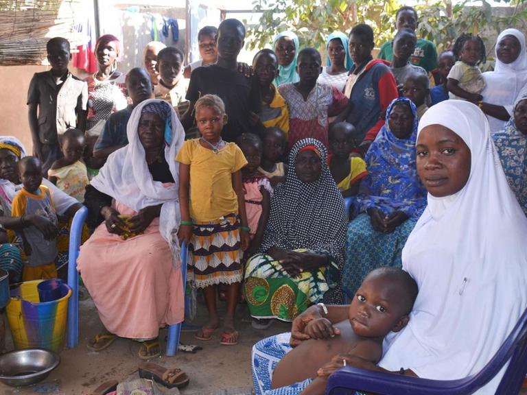 Fatimata Ourba sitzt mit ihren vier Kindern, anderen Familienmitgliedern und Nachbarn auf einem blauen Plastikstuhl und guckt in die Kamera. Sie ist geflüchtet aus der Sahel-Region, lebt jetzt aber in in Pazani - ein Viertel im Norden von Ouagaodougou.