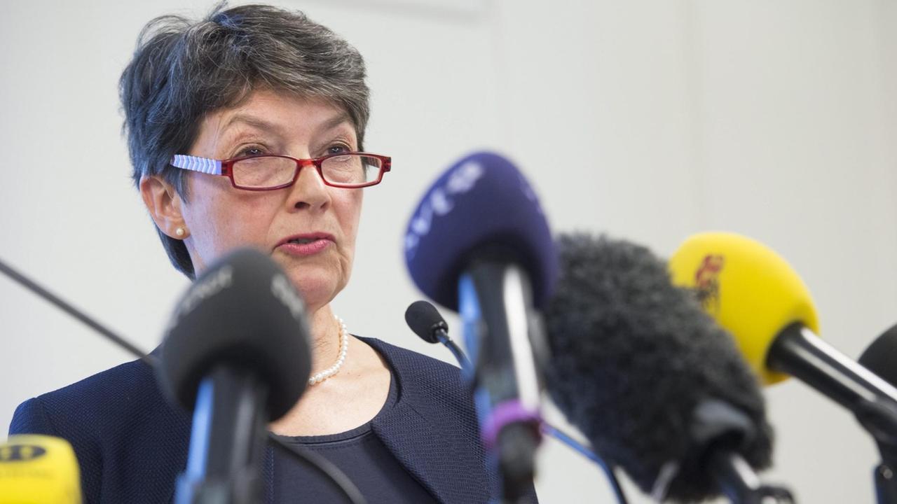 Ny trägt eine rotgerandete Brille und spricht mit ernstem Blick in mehrere Mikrofone. Ny trägt eine rotgerandete Brille und spricht mit ernstem Blick in mehrere Mikrofone.