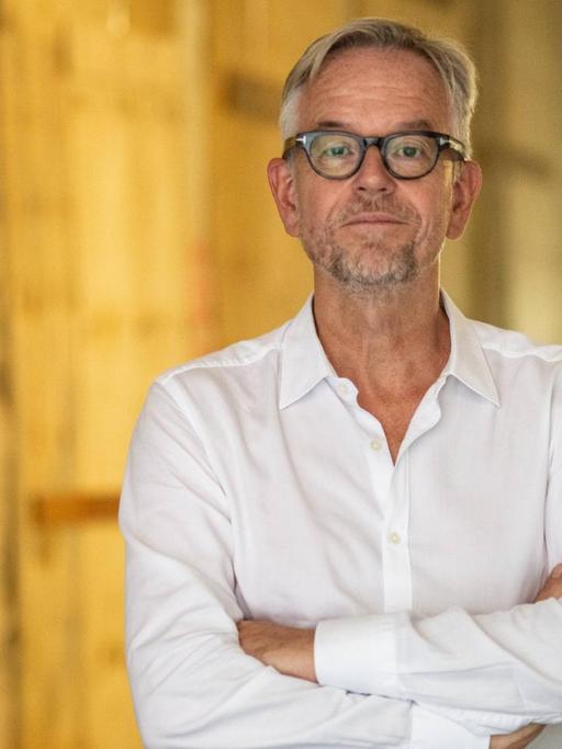 Porträt von Oliver Reese. Der Intendant des Berliner Ensemble steht vor der Baustelle des neuen Gebäudes. Er trägt ein weißes Hemd und hat die Arme vor der Brust verschränkt.