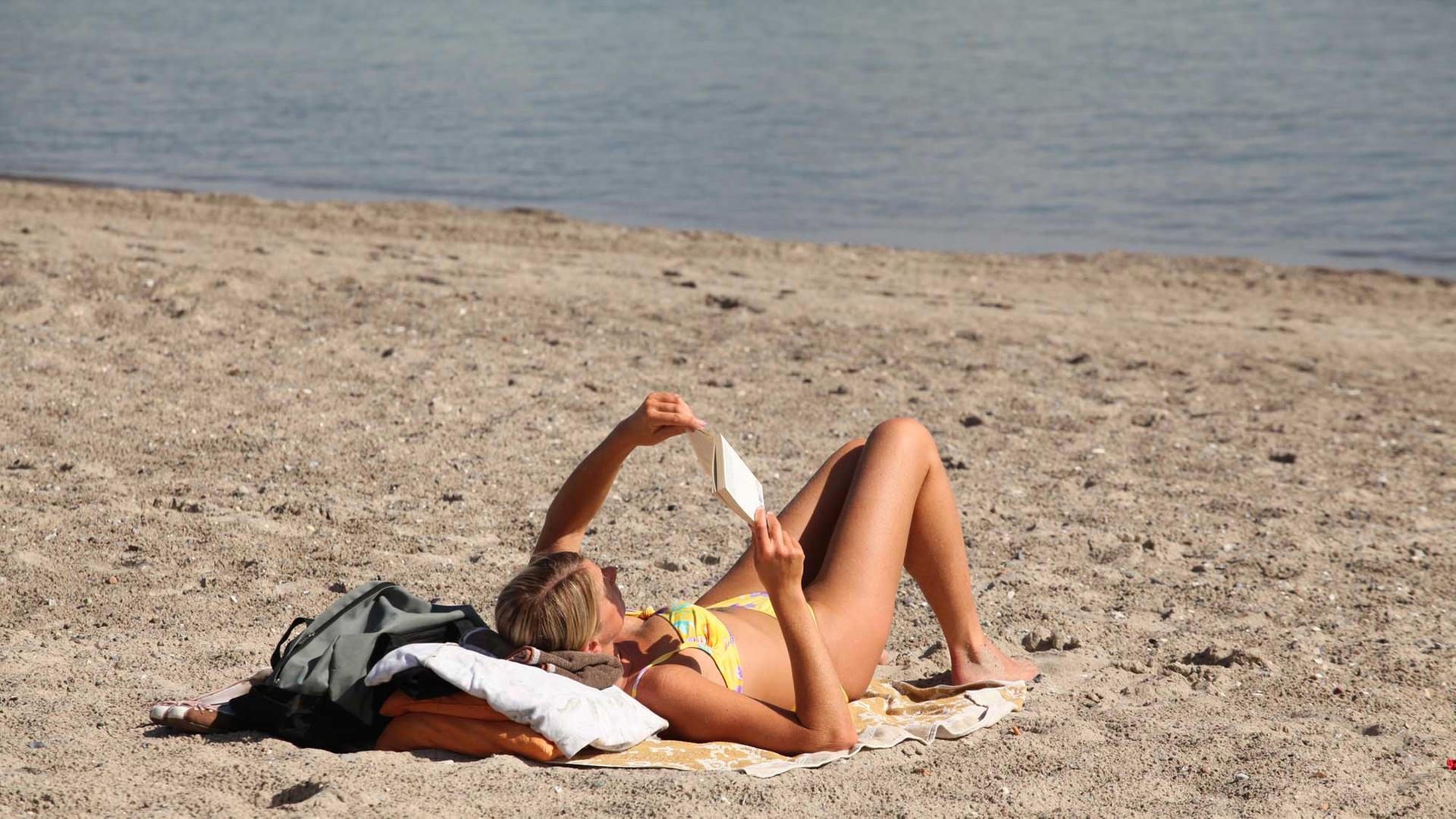 Eine Frau im Bikini liegt am Strand an der Ostsee und liest ein Buch.
