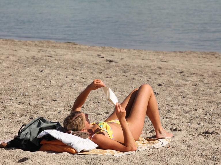 Eine Frau im Bikini liegt am Strand an der Ostsee und liest ein Buch.
