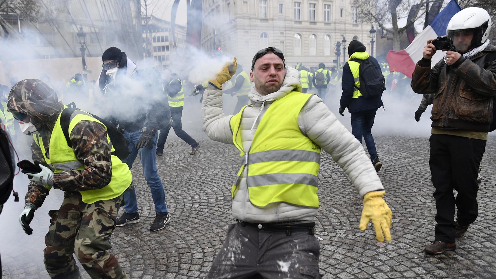 Ein Mann mit einer gelben Verkehrsweste steht auf der Straße und holt aus, um etwas nach vorn zu werfen. Er wird von einem anderen gefilmt. Es ist Rauch im Hintergrund. Ein Mann mit einer gelben Verkehrsweste steht auf der Straße und holt aus, um etwas nach vorn zu werfen. Er wird von einem anderen gefilmt. Es ist Rauch im Hintergrund.