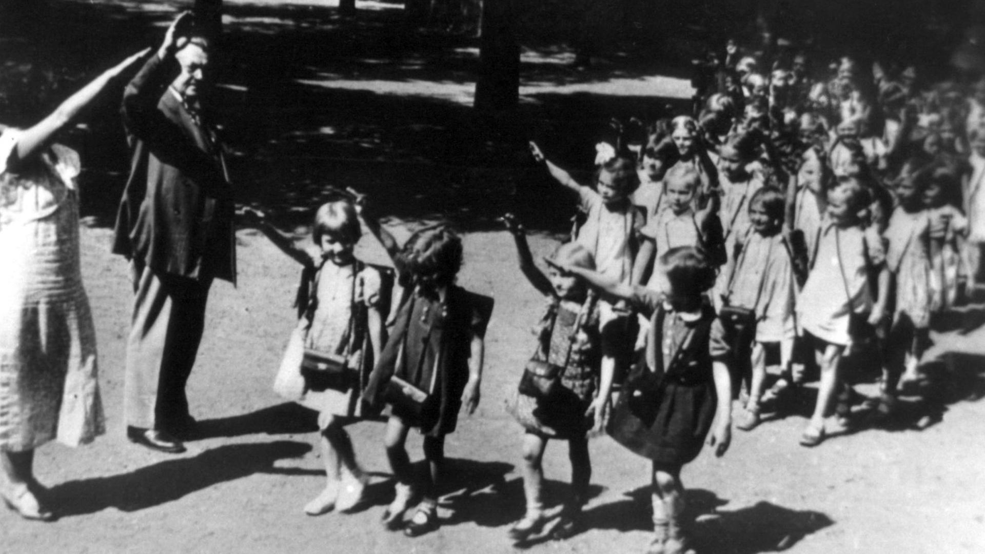 Die rechte Hand zum Hitlergruß erhoben gehen Erstklässler 1933 in Viererreihen an zwei ebenfalls grüßenden Erwachsenen (vermutlich Lehrpersonal) vorbei. Die rechte Hand zum Hitlergruß erhoben gehen Erstklässler 1933 in Viererreihen an zwei ebenfalls grüßenden Erwachsenen (vermutlich Lehrpersonal) vorbei.