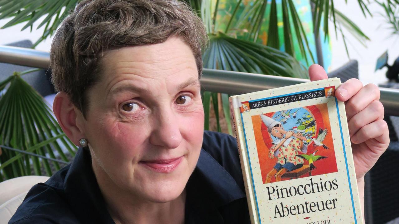 Die Autorin Felicitas Hoppe hält vor einer grünen Pflanze sitzend das Buch "Pinocchio" in die Kamera.