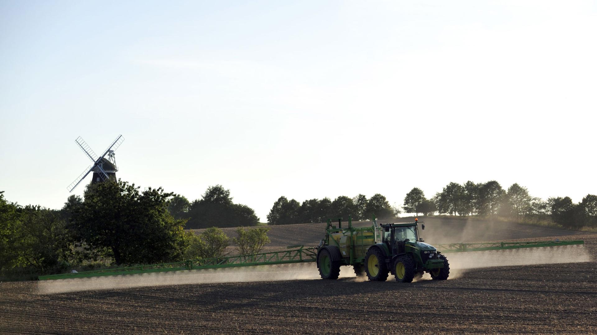 Ein Bauer besprüht mit einem Traktor sein Feld, im Hintergrund ist eine Mühle zu sehen.