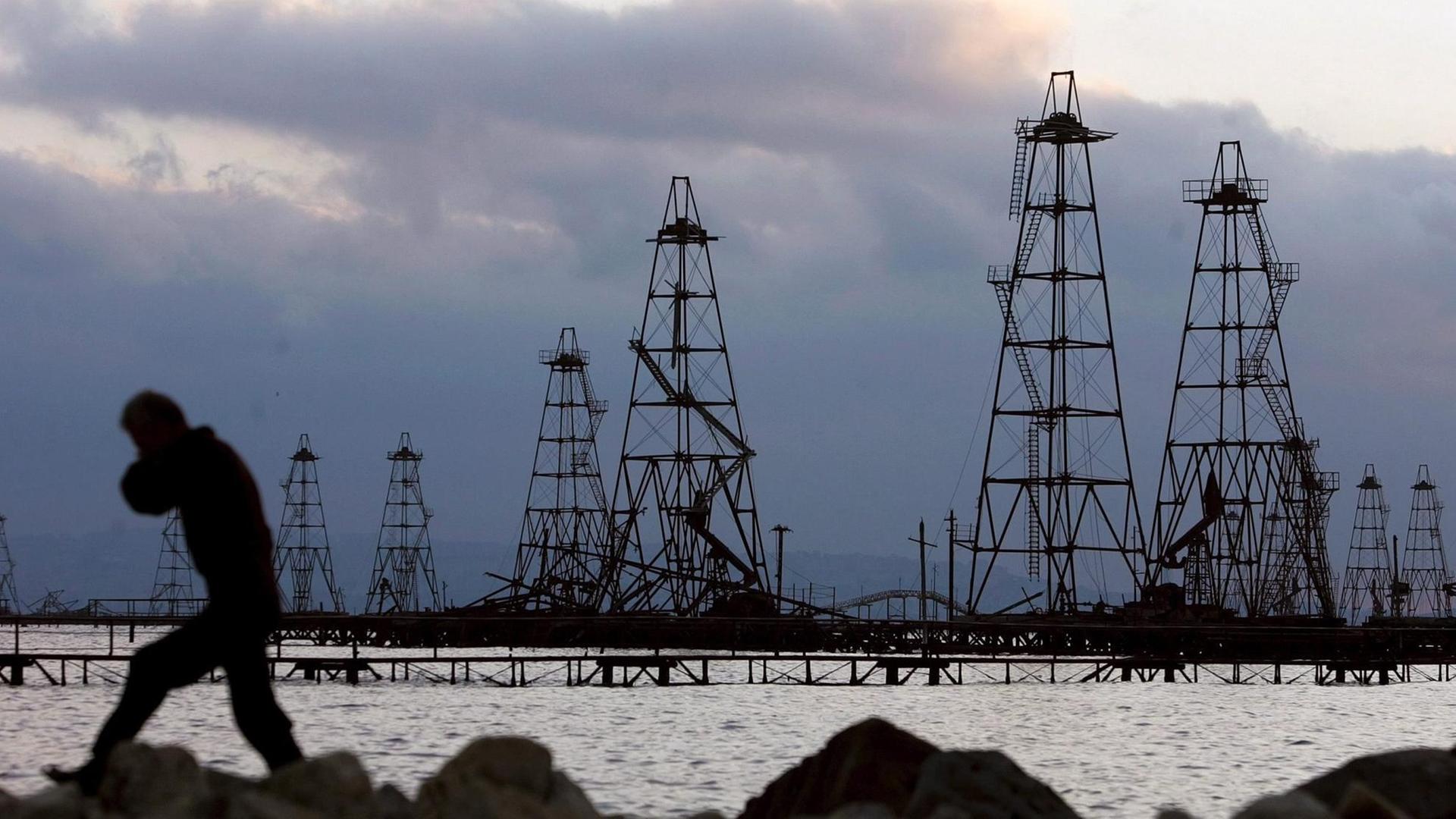 Ölbohrtürme im Kaspischen Meer in der Abenddämmerung. Davor geht am Ufer ein Mann entlang