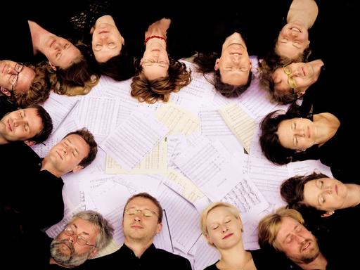 Mitglieder des Kammerchors Josquin de Préz aus Leipzig liegen mit geschlossenen Augen auf dem Boden, ihre Köpfen bilden einen Kreis und liegen auf Notenblättern.