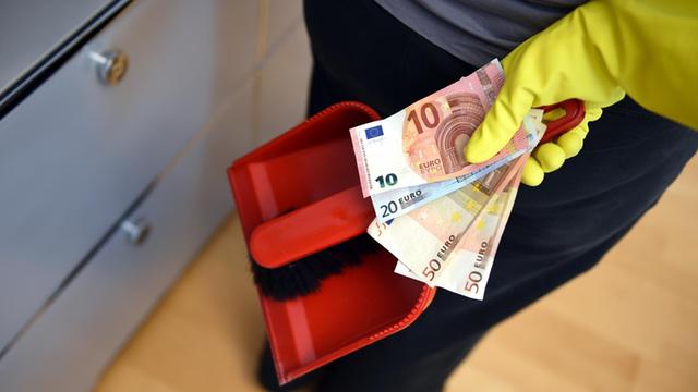 Eine Frau hält mehrere Geldscheine und einen Handfeger samt Kehrblech hinter ihrem Rücken.